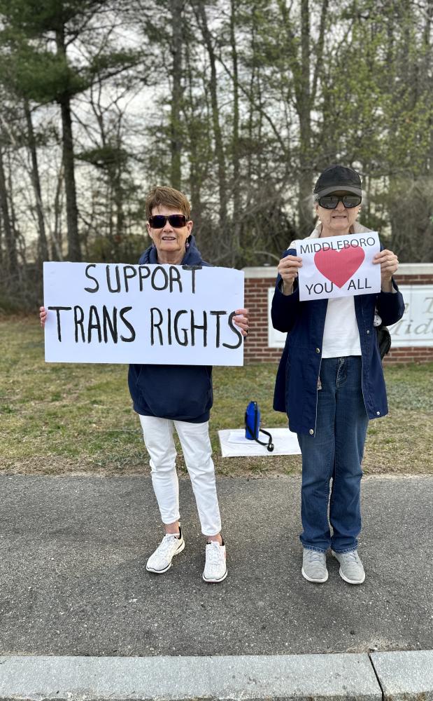 Two people stand in front of Nichols Middle School with signs saying “support trans rights” and “Middleboro loves you all.”
